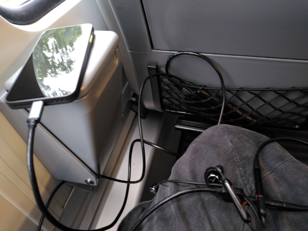 The back of a train seat. The black Fairphone Gen. 6 is placed ontop of a bin on the wall, with the bright yellow-greenish switch sticking out to the side. It is plugged into a cable leading to a power bank placed in the storage net in front. Another cable from that same powerbank leads to a pair of bluetooth headphones placed in the photographer's lap. The cables intermingle.